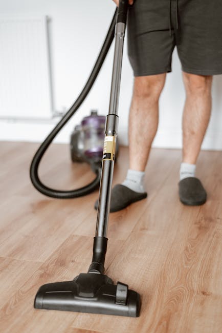 Close-up of a person using a handheld vacuum cleaner to perform surface cleaning on a light wooden floor in a residential setting. The individual is wearing dark shorts, grey socks, and grey slippers,