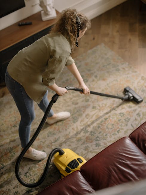 A person using a yellow and black vacuum cleaner to perform surface cleaning and deep cleaning on a patterned carpet in a domestic setting, with the vacuum's open top revealing a dust compartment, in