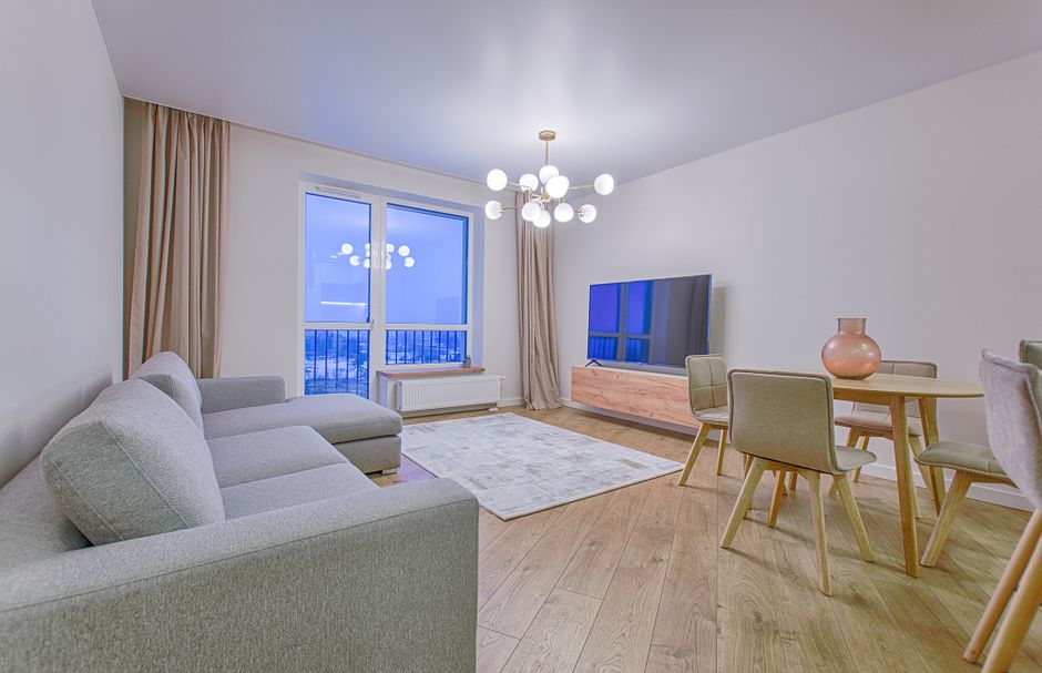 A modern, well-lit living room with light-colored walls and wooden flooring. The room features a large window with beige curtains, letting in natural light during daytime. There is a grey fabric sofa 